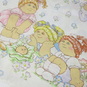 May include: White fabric featuring a repeating pattern of Cabbage Patch Kids dolls in various poses. The dolls have blonde, red, and light brown hair, wearing dresses and bows. The background includes blue and pink flowers and green grass.
