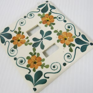 Può includere: Copri interruttore in ceramica decorativo con un design floreale. La copertura bianco sporco presenta fiori arancioni con foglie verdi e accenti turchese. Il design include volute e un motivo simmetrico.