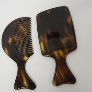 May include: A comb and a hand mirror, both featuring a tortoiseshell pattern in shades of brown and yellow. The comb has a curved handle and fine teeth. The mirror has a rectangular head and a curved handle, typical of hair styling tools.