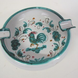 May include: White ceramic ashtray with a green and gold rooster design and floral accents. The ashtray has two side compartments for cigarettes.