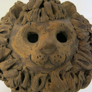 Puede incluir: Una escultura de cabeza de león de cerámica marrón con una melena texturizada y rasgos faciales detallados, incluidos los ojos, la nariz y los bigotes.