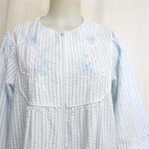 May include: A light blue and white striped robe with a white zipper closure. The robe features embroidered blue floral designs on the chest. The robe has long sleeves and a round neckline.