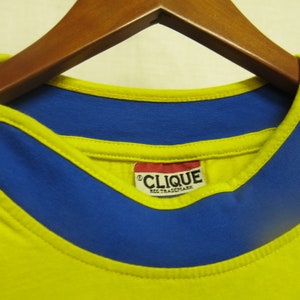 May include: Yellow t-shirt with a blue collar and a red tag that says "CLIQUE REG. TRADEMARK".