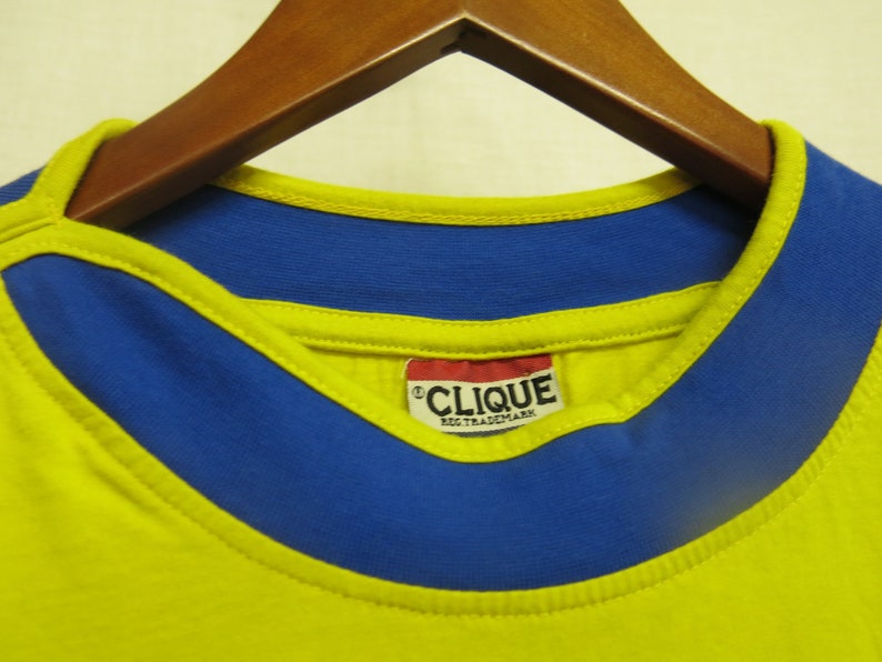 May include: Yellow t-shirt with a blue collar and a red tag that says "CLIQUE REG. TRADEMARK".