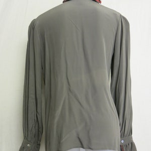Op de afbeelding: Een lang mouw, grijs blouse met een gerimpelde kraag en manchetten. De kraag en manchetten hebben een rode rand.
