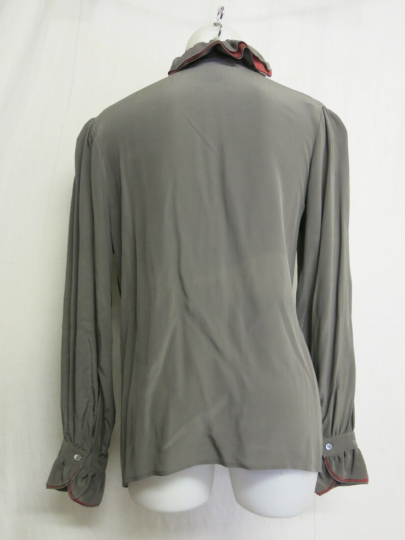 Op de afbeelding: Een lang mouw, grijs blouse met een gerimpelde kraag en manchetten. De kraag en manchetten hebben een rode rand.