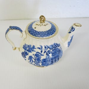Puede incluir: Tetera de cerámica color crema con estampado azul y blanco de Willow. La tetera tiene un cuerpo redondeado, un asa curva y un pico. La tapa está rematada con un adorno dorado. El asa y el pico tienen detalles dorados.