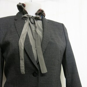 Op de afbeelding: Een donkergrijze blazer met een grijs en wit gepatroneerd overhemd eronder. Het overhemd heeft een grijze strik.