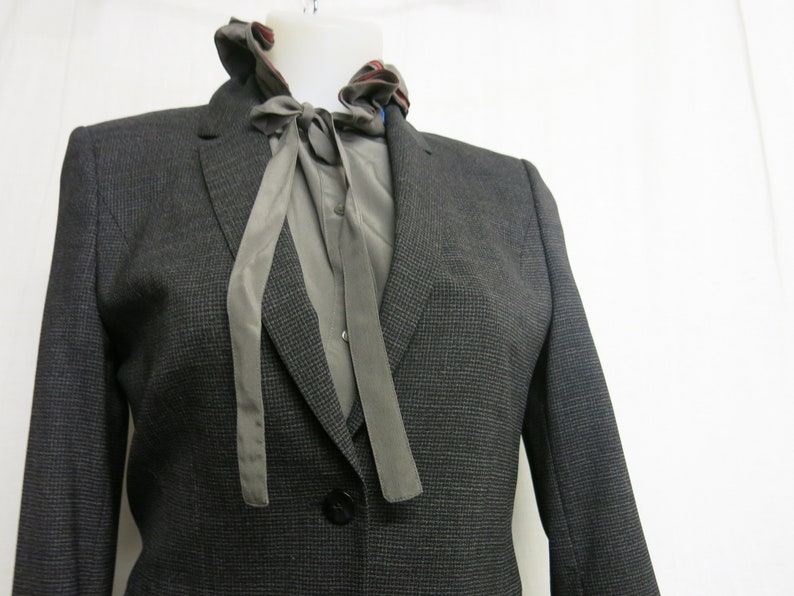 Op de afbeelding: Een donkergrijze blazer met een grijs en wit gepatroneerd overhemd eronder. Het overhemd heeft een grijze strik.