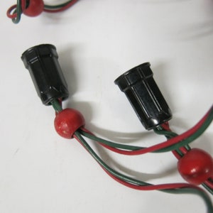May include: Close-up of a string of Christmas lights. The lights have black plastic sockets, red beads, and green and red wires. The background is white.