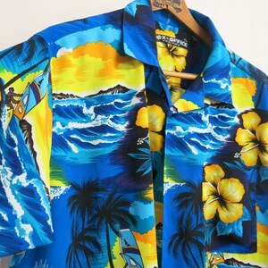 Pode incluir: Uma camisa havaiana azul e amarela com estampa de palmeira, onda do mar e flor de hibisco. A camisa tem colarinho abotoado e mangas curtas. O texto "EX-OFFICE" está impresso no colarinho.