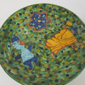May include: Hand-painted ceramic plate with a green background speckled with colorful dots. The plate features two figures and a star-shaped design in blue, orange, and yellow. The figures are in blue and yellow.