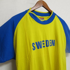 May include: Yellow and blue short-sleeved t-shirt with the word "SWEDEN" printed in blue on the front. The shirt has a blue and yellow color block design.