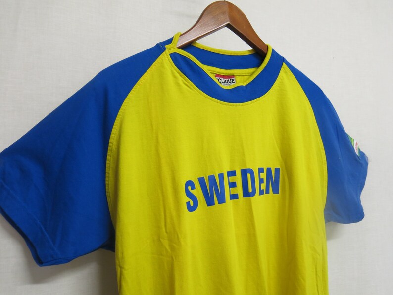 May include: Yellow and blue short-sleeved t-shirt with the word "SWEDEN" printed in blue on the front. The shirt has a blue and yellow color block design.