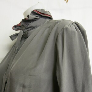 Op de afbeelding: Een grijs zijden blouse met een gerimpelde kraag en een rode bies. De blouse heeft lange mouwen en een strik aan de hals.