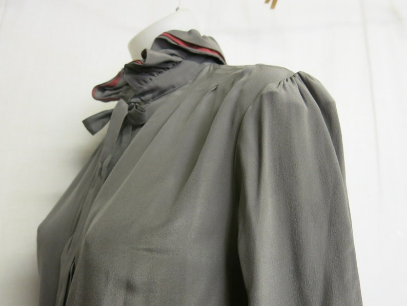 Op de afbeelding: Een grijs zijden blouse met een gerimpelde kraag en een rode bies. De blouse heeft lange mouwen en een strik aan de hals.