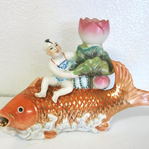 May include: A ceramic ornament featuring a child figure riding a large orange fish with gold detailing. The child wears blue and white clothing and holds a lotus flower. The fish is approximately 25 cm long. A decorative piece with a traditional design.