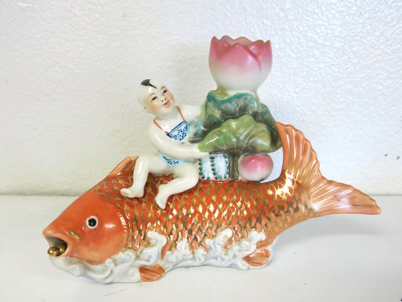 May include: A ceramic ornament featuring a child figure riding a large orange fish with gold detailing. The child wears blue and white clothing and holds a lotus flower. The fish is approximately 25 cm long. A decorative piece with a traditional design.