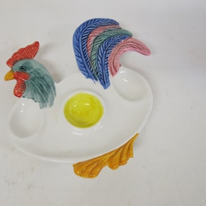 May include: A white ceramic rooster-shaped egg serving dish. The dish features a yellow yolk-shaped center, two egg cup holders, and a colorful rooster head and tail. The rooster's head is blue and green, with a red comb and yellow beak.