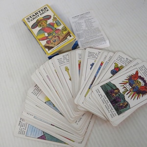 May include: A Starter Tarot Deck set featuring a yellow box with a colorful illustration of a figure, a white instruction booklet, and a fan of rectangular tarot cards with various illustrations and printed text descriptions on each card.