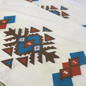 May include: White fabric with a repeating pattern of brown, blue, and red geometric shapes. The pattern is a stylized design of triangles, squares, and diamonds.