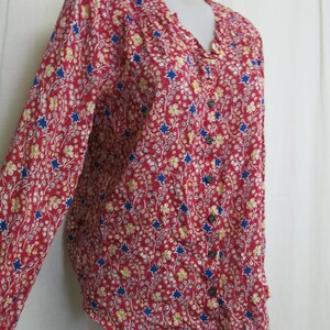 May include: A long-sleeved red button-up shirt with a floral and star print. The shirt has a ruffled collar and blue buttons.