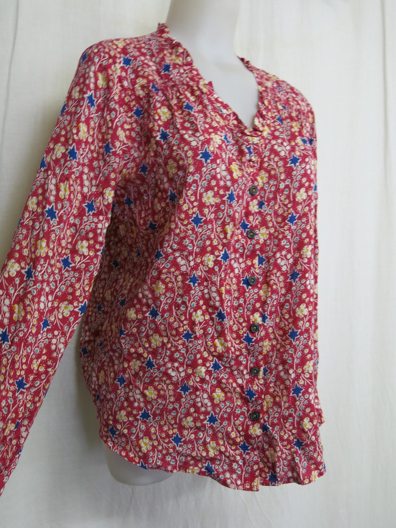 May include: A long-sleeved red button-up shirt with a floral and star print. The shirt has a ruffled collar and blue buttons.
