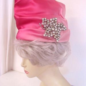 Op de afbeelding: Een felroze satijnen hoed met een zilverkleurige broche in de vorm van een blad, bezet met strass-steentjes. De hoed wordt getoond op een mannequinhoofd met lichtpaars haar, een vintage modeaccessoire.