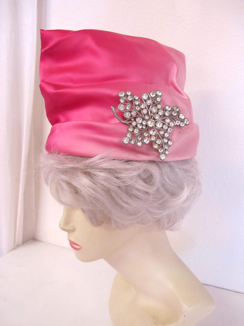 Op de afbeelding: Een felroze satijnen hoed met een zilverkleurige broche in de vorm van een blad, bezet met strass-steentjes. De hoed wordt getoond op een mannequinhoofd met lichtpaars haar, een vintage modeaccessoire.