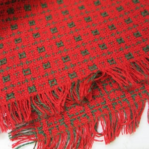 May include: Red and green woven fabric with a fringed edge. The fabric has a repeating pattern of small squares.