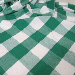 May include: A green and white checkered fabric, likely a tablecloth or similar textile. The pattern consists of large, evenly sized squares in a classic gingham design. The fabric is folded and draped, showing the texture and weave.