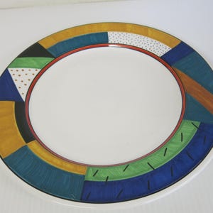 Op de afbeelding: Een witte keramische plaat met een kleurrijk geometrisch ontwerp rond de rand. Het ontwerp omvat secties in blauw, geel, groen en zwart, met een rode binnenring. De plaat heeft een moderne, artistieke uitstraling.