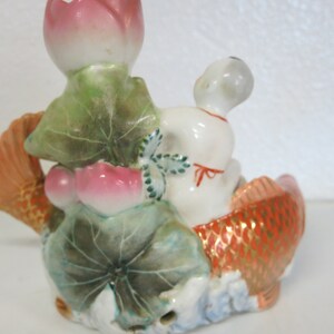 May include: A ceramic figurine of a white figure in a fish-shaped boat. The fish is orange with gold detailing. Pink lotus flowers and green leaves are also present, creating a decorative ornament.