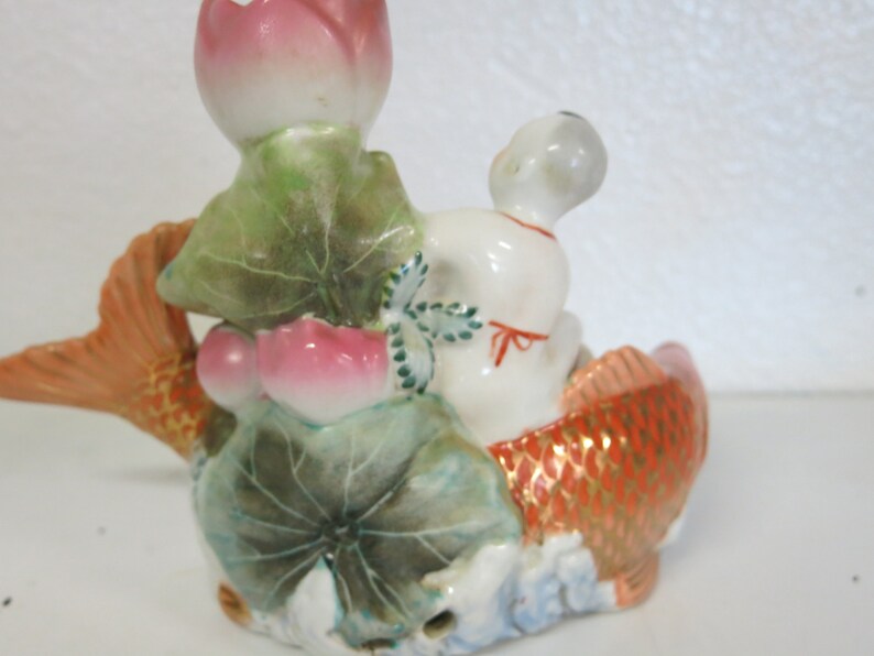 May include: A ceramic figurine of a white figure in a fish-shaped boat. The fish is orange with gold detailing. Pink lotus flowers and green leaves are also present, creating a decorative ornament.