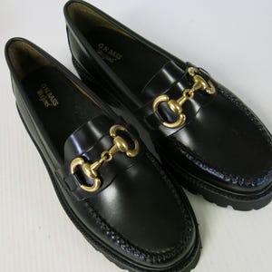 May include: Pair of black leather loafers with gold-colored horsebit hardware. The shoes are from the brand G.H. Bass Weejuns. The loafers have a classic design with a rounded toe and a stitched sole.