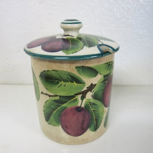 Könnte beinhalten: Keramiktopf mit Deckel, verziert mit einem handgemalten Design aus Pflaumen und grünen Blättern auf beigem Hintergrund. Der Deckel hat einen kleinen Knopf und eine grüne Umrandung. Küchenbehälter.