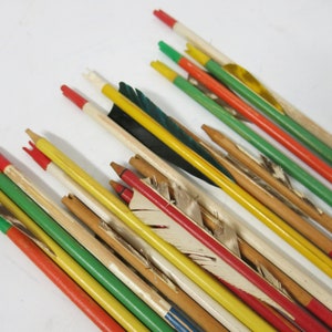 Puede incluir: Una colección de flechas de madera pintadas de rojo, amarillo, verde y blanco. Las flechas tienen plumas y están dispuestas en una pila.