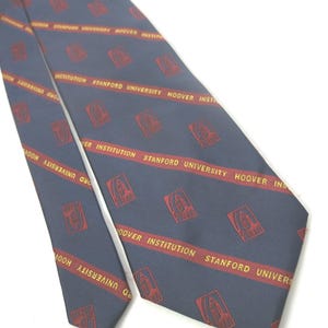 May include: A navy blue tie with a repeating pattern of red text and emblems. The text reads "STANFORD UNIVERSITY HOOVER INSTITUTION". The tie is angled across a white background.