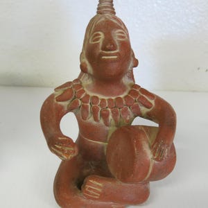 May include: A terracotta figurine of a seated figure holding a round object. The figure has a detailed face, a necklace, and a headdress. The sculpture is a reddish-brown color.