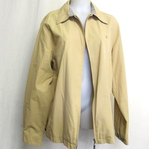 May include: Tan zip-up jacket with a collared neckline. The jacket has a small embroidered logo on the chest and a plaid pattern on the inside collar. The jacket is displayed on a mannequin against a white backdrop.