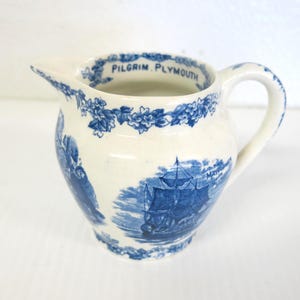 May include: A cream-colored ceramic creamer featuring blue transferware illustrations of a sailing ship and the text PILGRIM PLYMOUTH. The rim is decorated with a blue floral pattern, and the piece includes a curved handle and a pouring spout.