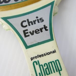 Puede incluir: Primer plano del mango de una raqueta de tenis vintage. El mango es blanquecino con detalles verdes y dorados. El nombre "Chris Evert" está impreso en negro sobre un fondo plateado, y la palabra "Champ" es verde.