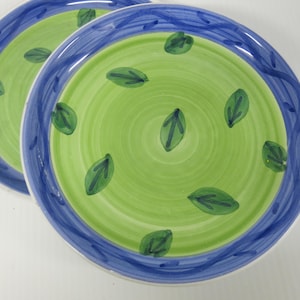 May include: Two ceramic plates with a green and blue design. The center of each plate is green with a spiral pattern and scattered green leaf designs. A blue border with a wavy pattern surrounds the green center.