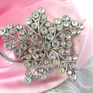Op de afbeelding: Een zilverkleurige broche in de vorm van een blad, versierd met talrijke heldere en lichtgele strass-steentjes. De broche is geplaatst tegen een achtergrond van roze satijn en witte vacht.
