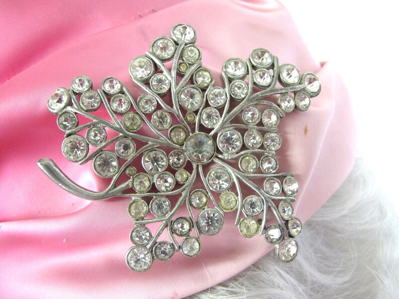 Op de afbeelding: Een zilverkleurige broche in de vorm van een blad, versierd met talrijke heldere en lichtgele strass-steentjes. De broche is geplaatst tegen een achtergrond van roze satijn en witte vacht.