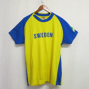May include: A yellow short-sleeved t-shirt with blue sleeves and a blue stripe down the sides. The shirt has the word "SWEDEN" printed in blue on the front.