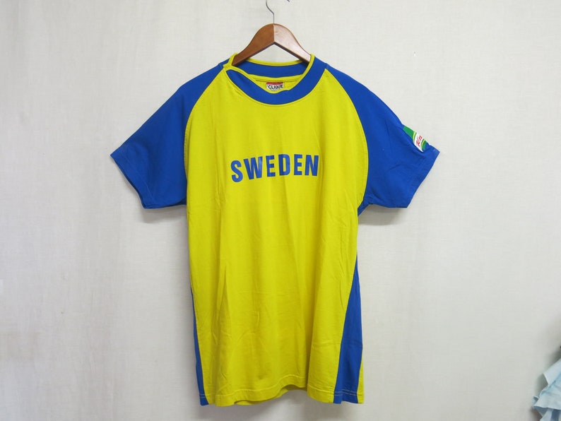 May include: A yellow short-sleeved t-shirt with blue sleeves and a blue stripe down the sides. The shirt has the word "SWEDEN" printed in blue on the front.