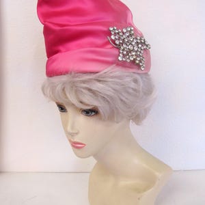 Op de afbeelding: Een felroze satijnen hoed met een zilverkleurige broche versierd met strass in een bloemmotief. De hoed heeft een afgeronde kroon en een contrasterende band. De hoed wordt getoond op een mannequinhoofd.
