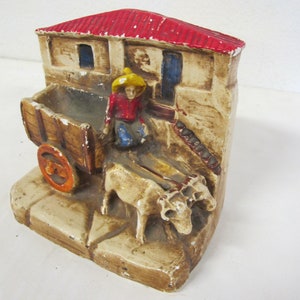 May include: A ceramic figurine depicting a rural scene with a red-roofed building, a wooden cart, and oxen. The figurine features a figure in a yellow hat and red shirt. The color palette includes beige, brown, red, and blue.