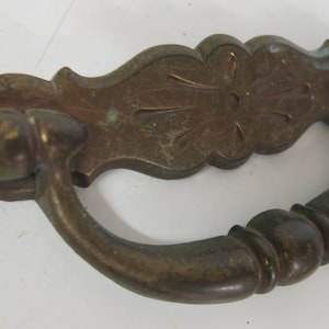 Puede incluir: Un tirador de puerta de latón vintage con un diseño ornamentado. El tirador tiene un mango curvo y una placa decorativa con un patrón floral.